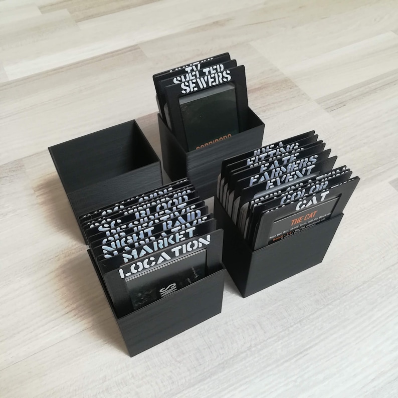 Puede incluir: Dos cajas negras llenas de peque&ntilde;as tarjetas negras. Las tarjetas tienen texto blanco, incluyendo "Night Raid", "Market", "Location", "Culi Tud Sewers", "Little Fate", "Farmers Event", "Color Colt" y "The Cat".