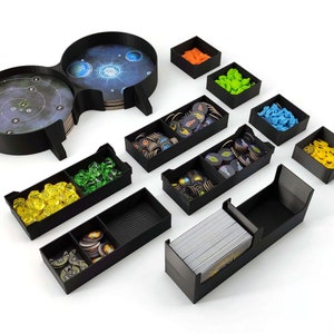Puede incluir: Un conjunto de organizadores de plástico negro para componentes de juegos de mesa. Los organizadores incluyen una bandeja de doble cara con un diseño de galaxia, una bandeja con un diseño de diana circular y varias bandejas más pequeñas para guardar diversas piezas de juego. Las piezas incluyen fichas amarillas, verdes, azules, naranjas y marrones.