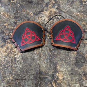 May include: A pair of black leather arm guards with red leather Celtic knot designs. The guards are laced together with black laces.