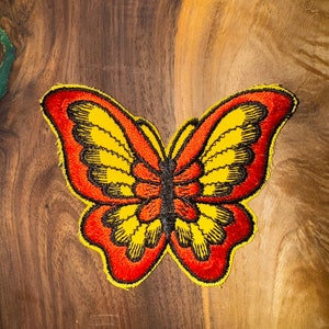 Puede incluir: Un parche bordado de mariposa rojo y amarillo con detalles negros. El parche está sobre una superficie de madera marrón.