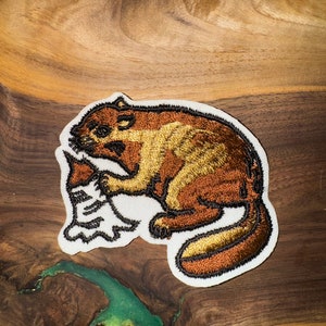 Pode incluir: Um remendo bordado marrom e dourado com um castor segurando um galho em suas patas. O remendo está sobre uma superfície de madeira.