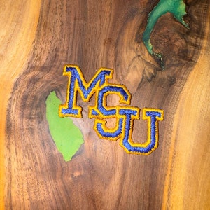 Op de afbeelding: Een houten plank met een groene harsinleg en een blauw en gouden geborduurd patch met de tekst "MSU".