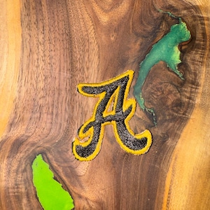 Könnte beinhalten: Ein gestickter Buchstabe "A" in Schwarz und Gelb auf einer Holzoberfläche mit grünen Harzakzenten.
