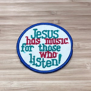 Könnte beinhalten: Ein runder weißer Patch mit blauem Rand. Der Patch hat den Text "Jesus has music for those who listen!" in roter, grüner und blauer Farbe gestickt.