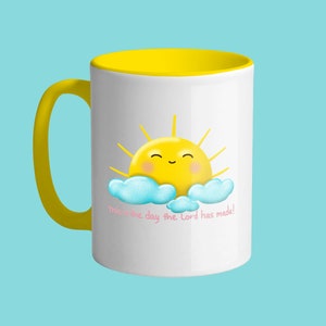 Könnte beinhalten: Weißer Keramikbecher mit gelbem Innenraum und Griff. Der Becher zeigt eine lächelnde gelbe Sonne mit blauen Wolken und dem Text "This is the day the Lord has made!" in Rosa.