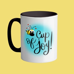 Könnte beinhalten: Weiße Keramiktasse mit schwarzem Rand und Henkel. Die Tasse hat einen blauen und weißen Ombre-Hintergrund mit einer Cartoon-Biene und dem Text "cup of joy!" in Schwarz.