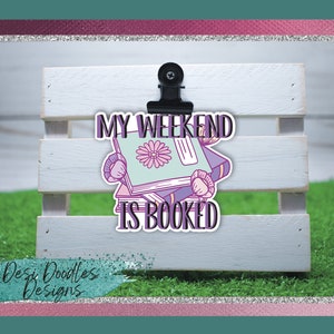Op de afbeelding: Een sticker met een stapel boeken en de tekst "My Weekend Is Booked". De boeken zijn paars, blauw en groen met een bloem op de voorkant van het bovenste boek. De sticker zit op een wit houten oppervlak.
