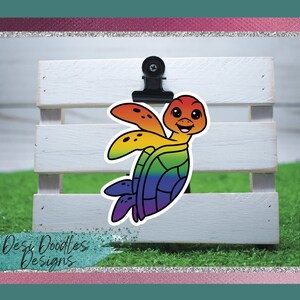 May include: A rainbow colored turtle sticker with a smiling face. The turtle is in a swimming pose with its flippers outstretched. The sticker is on a white wooden surface.