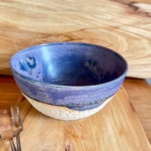 Könnte beinhalten: Eine handgefertigte Keramikschale mit tiefblauem Innenraum und strukturierter, cremefarbener Außenseite. Die Schale hat ein einzigartiges, rustikales Design mit einem Band dunkler Akzente am Rand. Silberne Servierutensilien sind im Vordergrund.