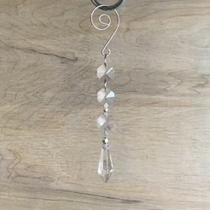 May include: A clear crystal chandelier ornament with a silver-colored metal hook. The ornament features a series of faceted crystal beads and a teardrop-shaped crystal pendant. The crystals reflect light, creating a sparkling effect.