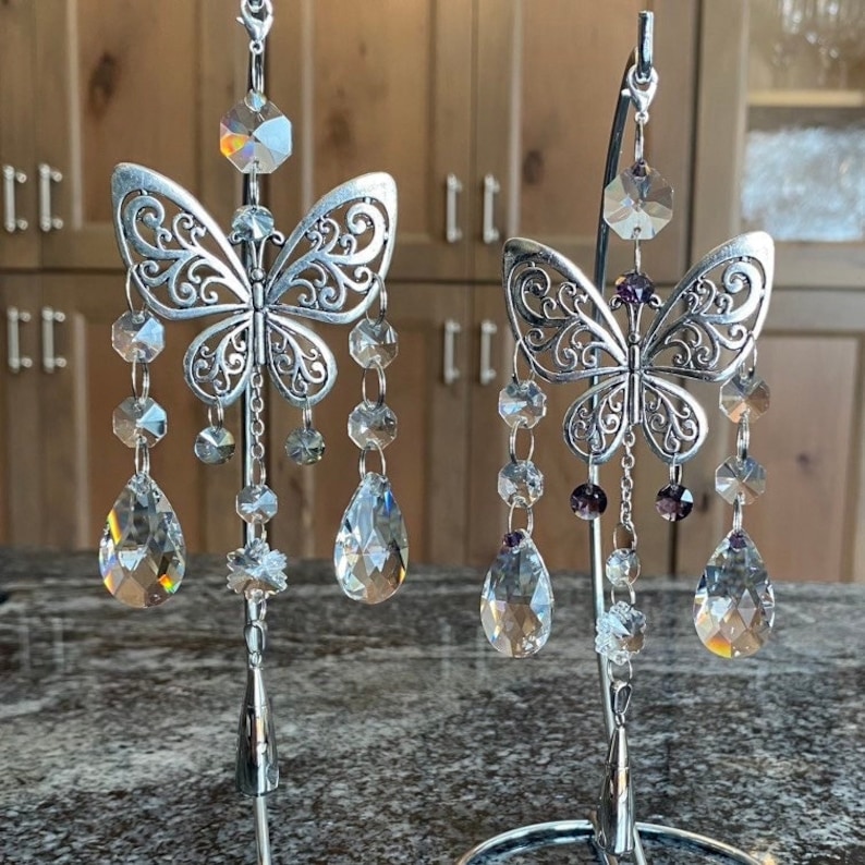 May include: Two silver butterfly-shaped crystal suncatchers hanging from a metal stand. Each butterfly has a crystal teardrop hanging from its wings. The butterfly on the left has clear crystals, while the butterfly on the right has clear and purple crystals.