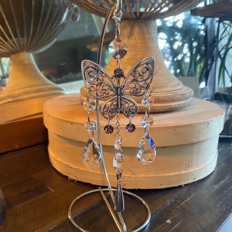 May include: A silver butterfly-shaped crystal suncatcher with iridescent crystals hanging from it. The suncatcher is on a silver stand.