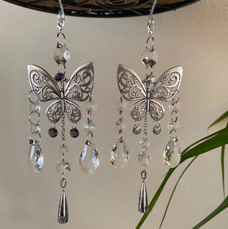 May include: A pair of silver butterfly earrings with crystal and purple bead accents. The earrings dangle from a silver chain and feature a delicate butterfly design.
