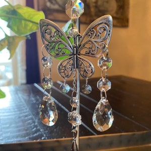 May include: A silver butterfly charm with crystal beads hanging from it. The butterfly has a filigree design and is attached to a metal rod.