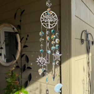 Puede incluir: Un carillón decorativo con un diseño de árbol de la vida plateado, adornado con cuentas de cristal iridiscentes, amuletos de sol, estrella y luna. El carillón incluye una pequeña campana en la parte inferior, creando una estética caprichosa y celestial.