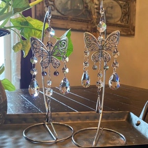 May include: Two silver butterfly-shaped crystal suncatchers hanging from a metal stand. Each suncatcher features a detailed butterfly design with dangling crystal beads and teardrop-shaped crystals.