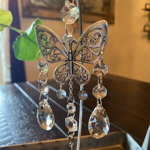 May include: A silver butterfly-shaped ornament with crystal beads hanging from it. The ornament is hanging from a metal stand.