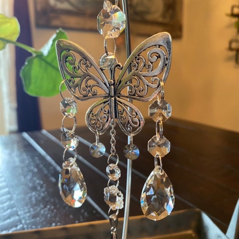 May include: A silver butterfly-shaped ornament with crystal beads hanging from it. The ornament is hanging from a metal stand.