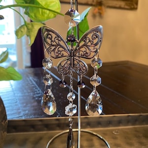May include: A silver butterfly-shaped crystal suncatcher with clear and purple crystals hanging from it. The suncatcher is hanging from a silver stand.