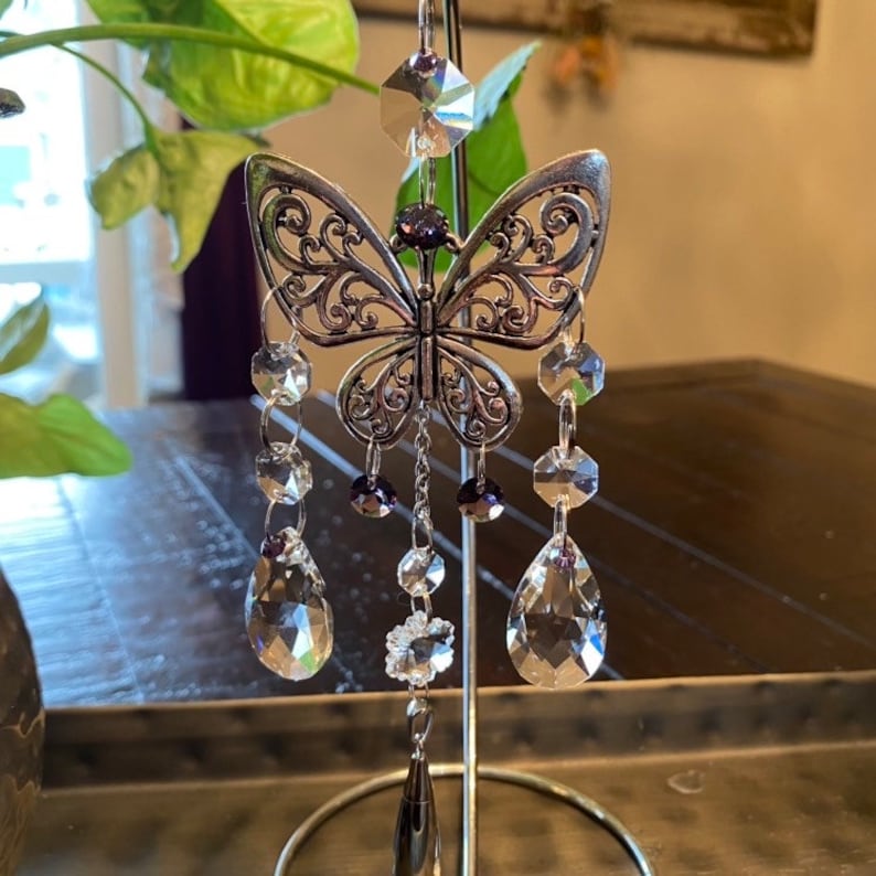 May include: A silver butterfly-shaped crystal suncatcher with clear and purple crystals hanging from it. The suncatcher is hanging from a silver stand.