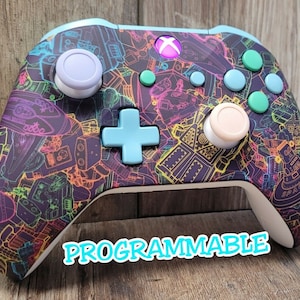 May include: A custom Xbox controller with a vibrant, space-themed design. The controller features a purple base with a colorful pattern of rockets, robots, and other space-related graphics. The buttons and thumbsticks are various colors, and the text "PROGRAMMABLE" is displayed.