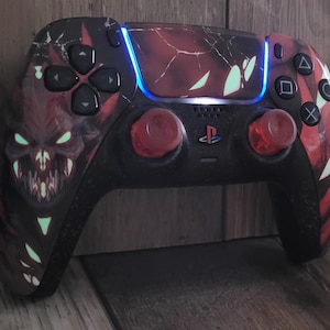 May include: Customized black and red PlayStation 5 controller with a demonic design. The controller features a red and black color scheme with a glowing green monster face on the left side. The thumbsticks are red, and the buttons are black.