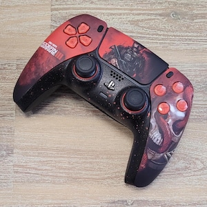 May include: A custom PlayStation 5 controller with a red and black design. The controller features a skull and snake graphic, along with the text "Call of Duty Modern Warfare III". The buttons are red.