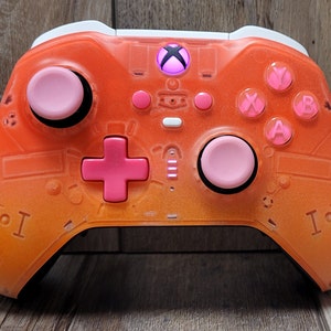 May include: An orange and pink Xbox controller with a transparent design. The controller features pink buttons, thumbsticks, and a directional pad. The Xbox logo is illuminated in the center. The controller is set against a wooden surface.