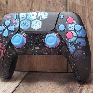May include: A black PlayStation 5 DualSense controller with a blue and pink water-drop design. The controller has a blue and pink button set.
