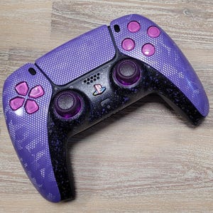 May include: A custom purple and black PlayStation 5 controller. The controller features a textured purple finish with pink accent buttons. The design includes a PlayStation logo and two analog sticks.