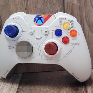 May include: A white Xbox controller with colorful buttons and a rainbow stripe design. The controller has a variety of labels, including "SCAN", "THROT", "FCS", "TOY", "MODE", "LOCK", and "FLT CTRL".