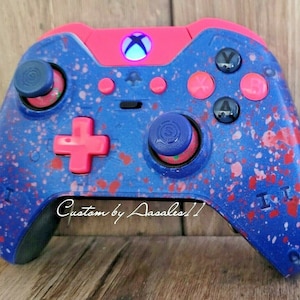 May include: Customized Xbox controller in blue with red and pink accents. The controller features a splatter paint design, pink buttons, and blue thumbsticks. The text "Custom by Aasales11" is visible on the controller.