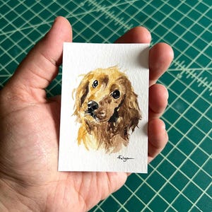 以下が含まれることがあります： 茶色とタン色の毛並みと暗い目をした犬の頭の水彩画。この作品は、小さな長方形の白い紙の上にあります。アーティストのサイン「Fulya」が右下にあります。