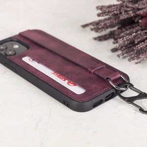 Puede incluir: Una funda de tel&eacute;fono de color morado oscuro con acabado texturizado, que incluye una ranura para tarjetas con una tarjeta blanca con texto en rojo, una correa horizontal y un mosquet&oacute;n de metal negro unido a una anilla en el lateral.