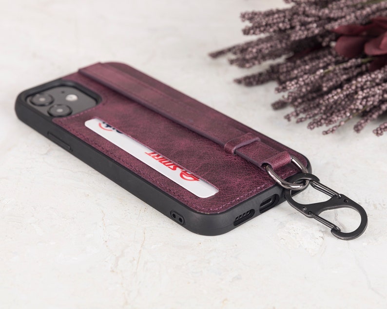 Puede incluir: Una funda de tel&eacute;fono de color morado oscuro con acabado texturizado, que incluye una ranura para tarjetas con una tarjeta blanca con texto en rojo, una correa horizontal y un mosquet&oacute;n de metal negro unido a una anilla en el lateral.
