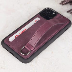 Puede incluir: Una funda de tel&eacute;fono de color morado oscuro con marco negro, que incluye una ranura para tarjetas con una tarjeta blanca con texto en rojo y una correa de cuero morada a juego sujeta a una anilla en D plateada. Abertura para triple c&aacute;mara.