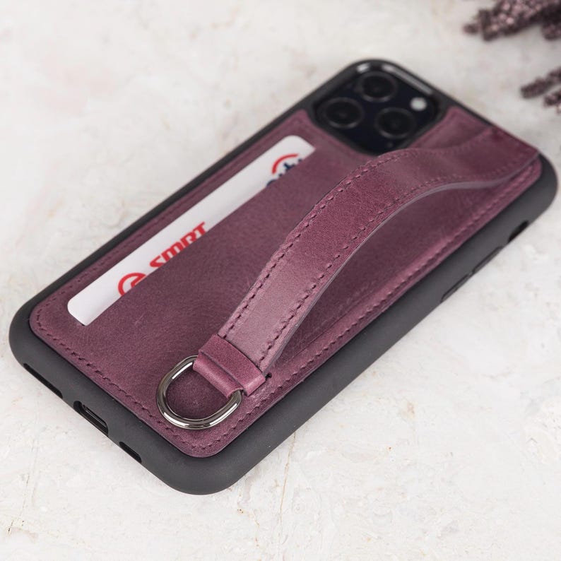 Puede incluir: Una funda de tel&eacute;fono de color morado oscuro con marco negro, que incluye una ranura para tarjetas con una tarjeta blanca con texto en rojo y una correa de cuero morada a juego sujeta a una anilla en D plateada. Abertura para triple c&aacute;mara.