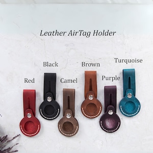 Könnte beinhalten: Leder-AirTag-Halter in verschiedenen Farben, darunter Schwarz, Braun, Türkis, Rot, Camel und Lila. Jeder Halter hat einen genähten Kreis für den AirTag.