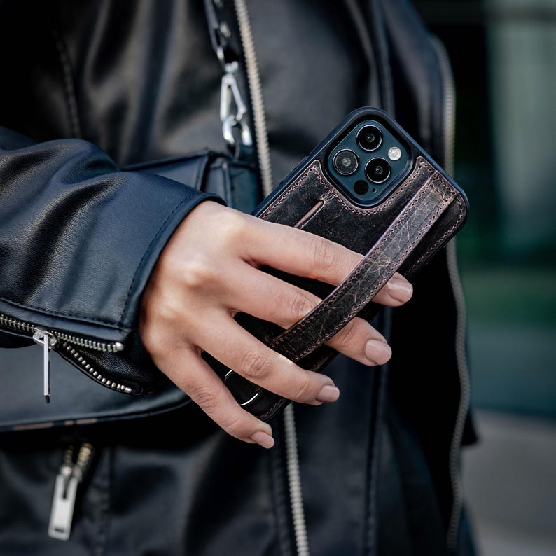 Puede incluir: Una funda de tel&eacute;fono de cuero envejecido en color marr&oacute;n oscuro con una correa vertical integrada en la parte posterior para mejorar el agarre. La funda tiene un recorte para las lentes de la c&aacute;mara y costuras visibles en los bordes de la correa.