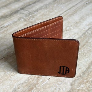 May include: A brown leather wallet with a bi-fold design. The wallet has a smooth texture and features the initials "JIP" in black on the front. The interior reveals multiple card slots and a contrasting light brown lining, with visible stitching.
