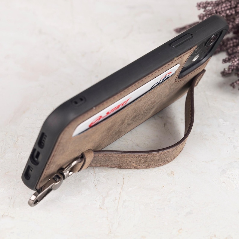 Puede incluir: Una funda de tel&eacute;fono de cuero marr&oacute;n con marco negro, que incluye una ranura para tarjetas con una tarjeta blanca con texto en rojo y azul, y una correa de mu&ntilde;eca de cuero marr&oacute;n desmontable sujeta con un clip de metal plateado en el lateral.