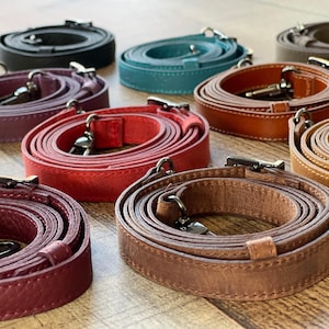 Puede incluir: Una colección de correas de cuero para bolsos enrolladas en varios colores, incluyendo negro, verde azulado, rojo, burdeos y varios tonos de marrón. Cada correa cuenta con mosquetones giratorios de metal y una trabilla de cuero deslizante.
