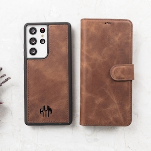 May include: A brown leather phone case with a black frame and a cutout for a multi-lens camera system, featuring a monogram with the letters KYM. Beside it is a matching brown leather wallet-style phone case with a magnetic closure strap.