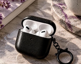 May include: A black textured protective case for wireless earbuds, shown in an open position with white earbuds inside. The case features a black circular keychain ring attached to the side, providing a secure way to carry the device while travelling.