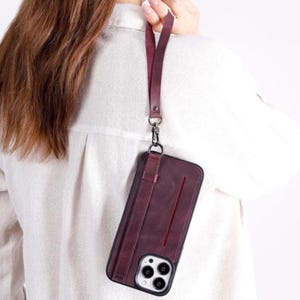 Puede incluir: Una funda de tel&eacute;fono color burdeos oscuro con una ranura vertical para tarjetas y una correa de mu&ntilde;eca desmontable a juego. La funda tiene un acabado mate, un marco negro alrededor de las lentes de la c&aacute;mara y un cierre met&aacute;lico plateado.