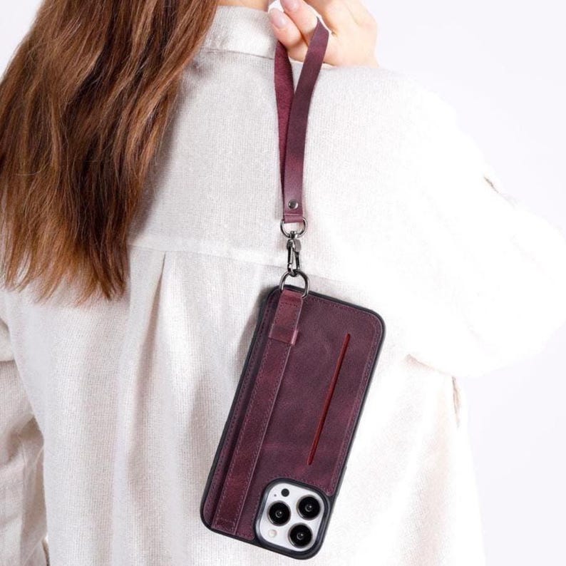 Puede incluir: Una funda de tel&eacute;fono color burdeos oscuro con una ranura vertical para tarjetas y una correa de mu&ntilde;eca desmontable a juego. La funda tiene un acabado mate, un marco negro alrededor de las lentes de la c&aacute;mara y un cierre met&aacute;lico plateado.