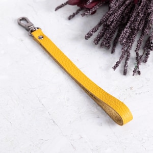 May include: A yellow leather wrist strap with a silver-colored clasp. The strap is a vibrant mustard color and has a textured surface. The background includes a sprig of dark purple flowers.
