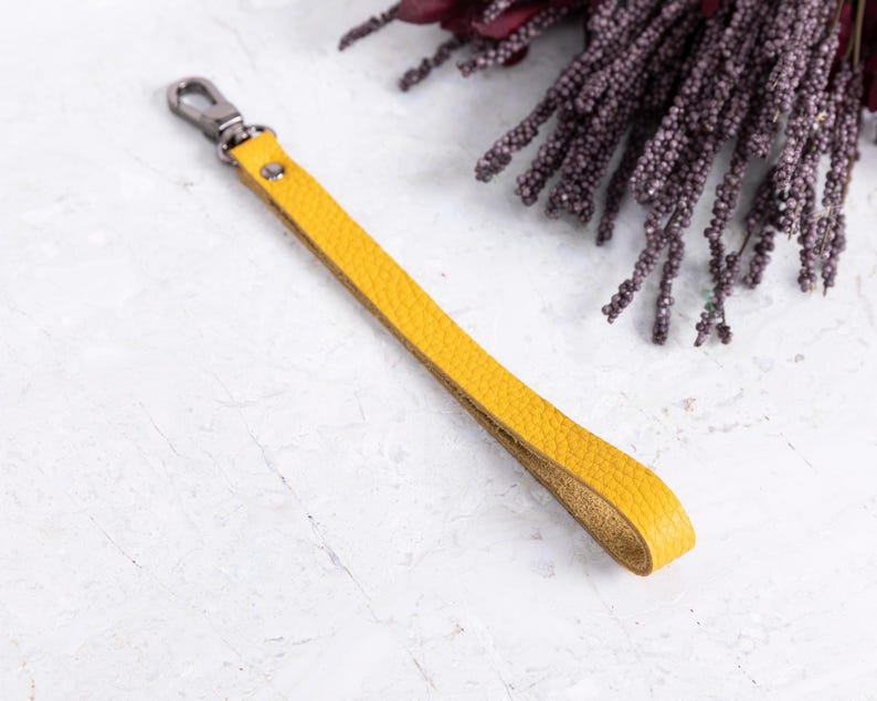 May include: A yellow leather wrist strap with a silver-colored clasp. The strap is a vibrant mustard color and has a textured surface. The background includes a sprig of dark purple flowers.
