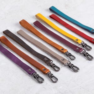 May include: A selection of leather wrist straps in a range of colours, such as purple, brown, yellow, and red. Each strap has a metal clasp for attachment. The straps are arranged diagonally against a light background.