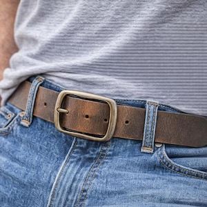 May include: A brown leather belt with a distressed finish and a rectangular brass-toned metal buckle. The belt is threaded through the belt loops of blue denim jeans, worn with a light gray textured t-shirt tucked into the waistband.